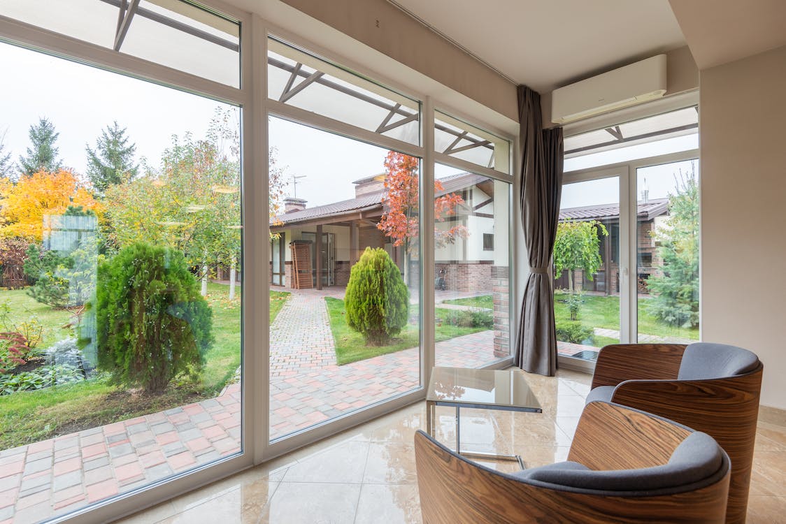 A view of a lush backyard from inside of a living room. Two chairs face a wall of three large windows. Beyond the windows are decorative paving and assorted shrubbery and plants. Another building can be seen beyond the green yard.