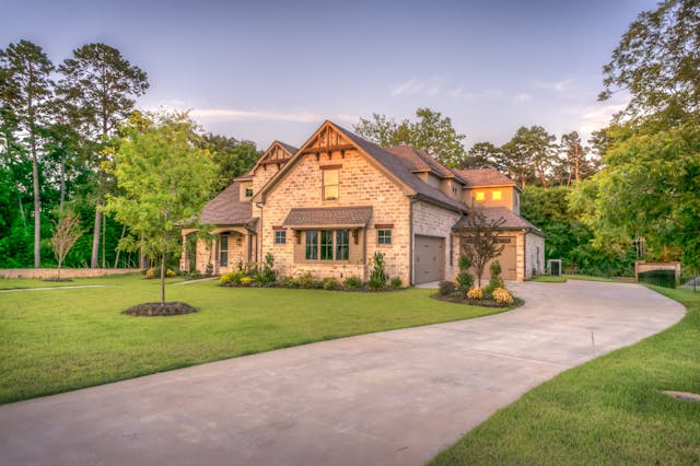 A photo of a tan brick two story house at the end of a long paved driveway.It is surrounded by lush green trees and a yard.