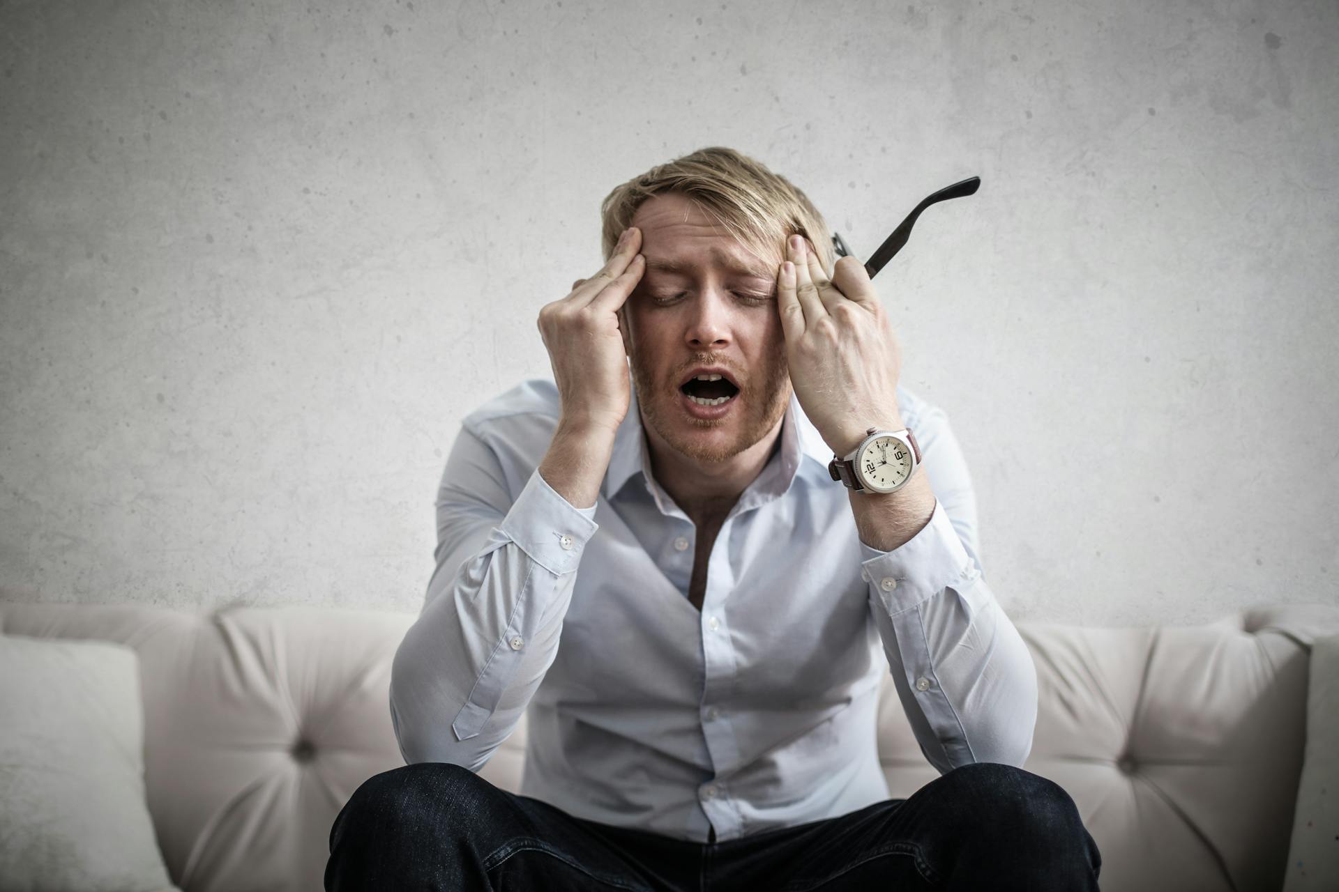 A man sits on a white couch with his hands pressed against the sides of his forehead. A look of agony is on his face, his eyes are closed and his mouth is open.