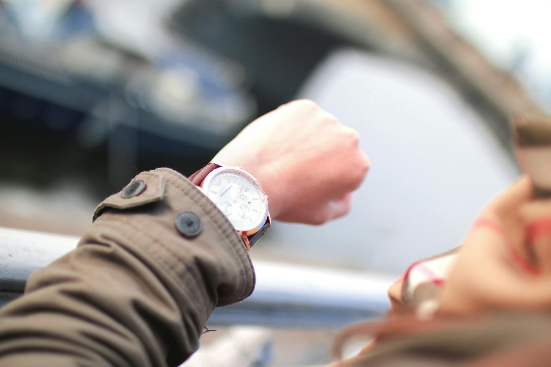 A close up photo of a hand and wrist. A person is checking their watch. The arm has a brown jacket on it, and the watch has a broad face and deep red band.