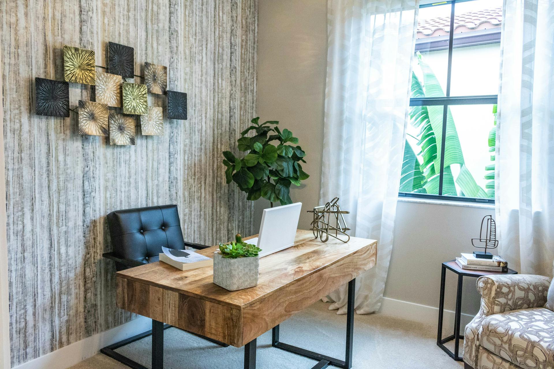A clean home office. There is a wooden desk with a laptop and desk decorations in front of a black leather chair. A potted plant and cozy chair decorate the room. A window provides sufficient lighting to the desk with a view of foliage outside of the window.