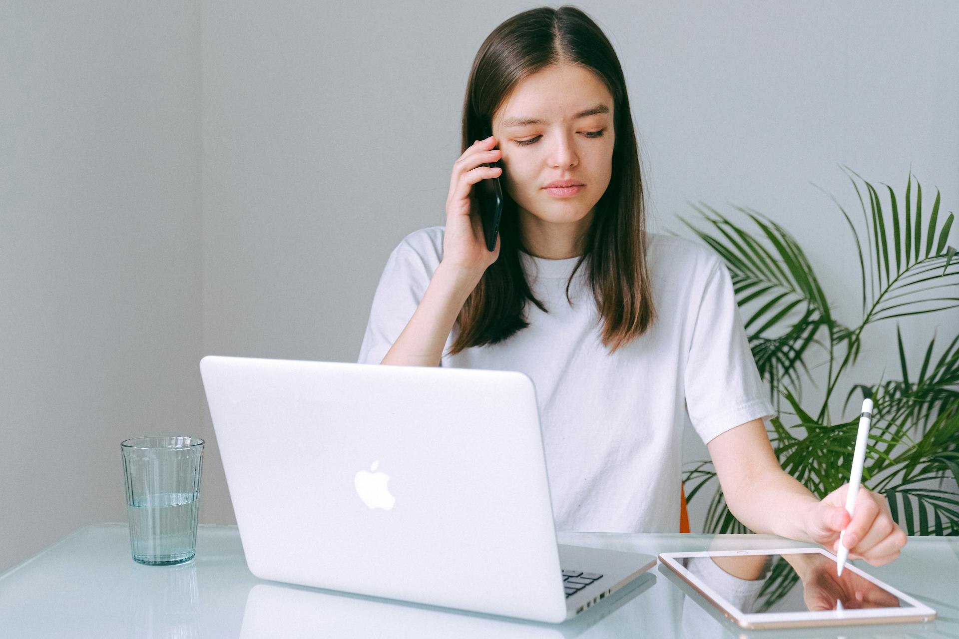 A young woman in a white T-shirt sits at a white glass desk. She holds a phone to her ear in one hand, and uses a stylus in her other hand to use a tablet resting on the desk. In front of her on the desk are also a laptop and a half full glass of water.