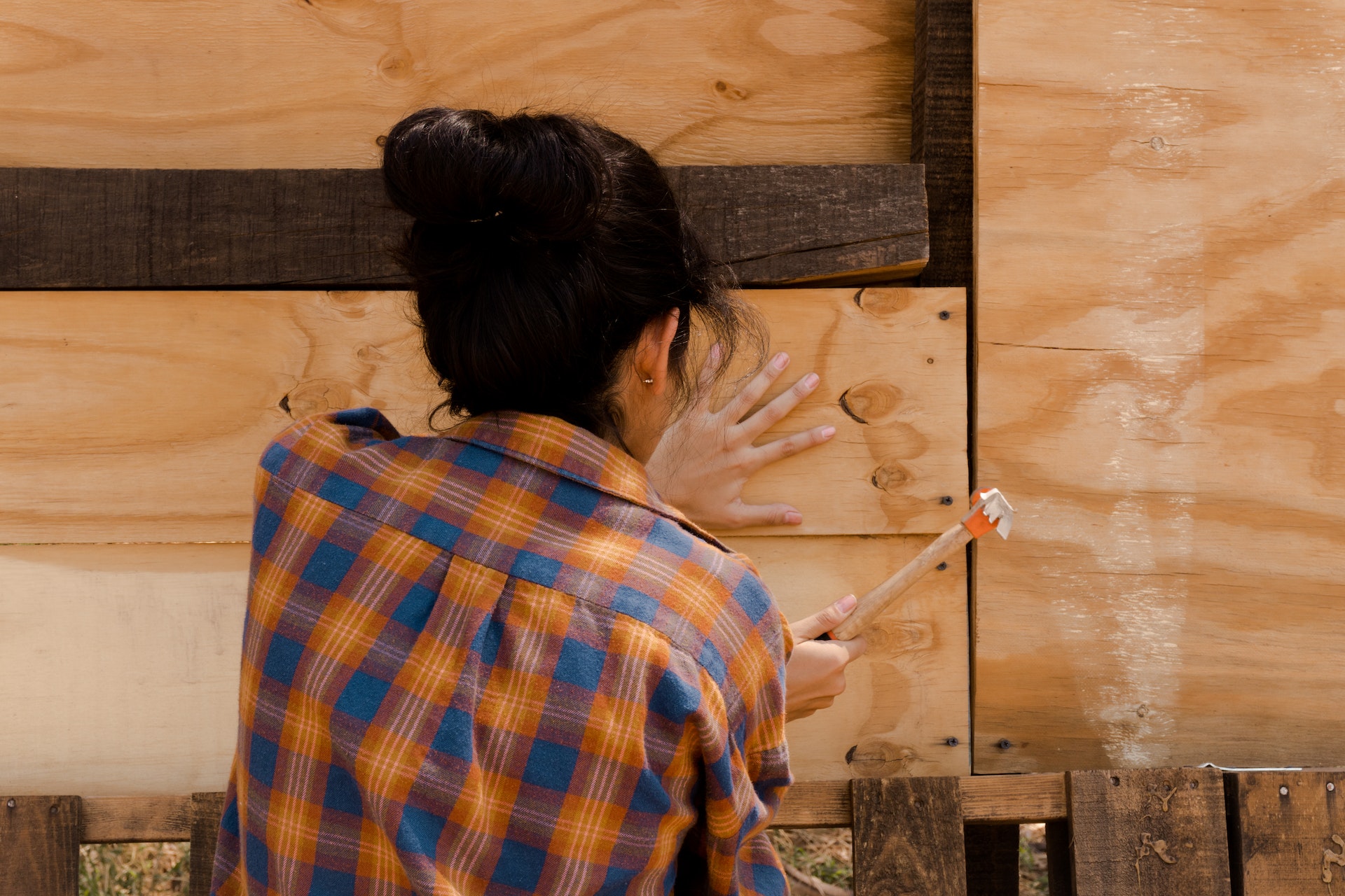 A view of a person from behind as they hammer nails into wooden plank fencing. The person has dark hair pulled up into a bun, and is wearing a blue and orange flannel patterned work shirt.