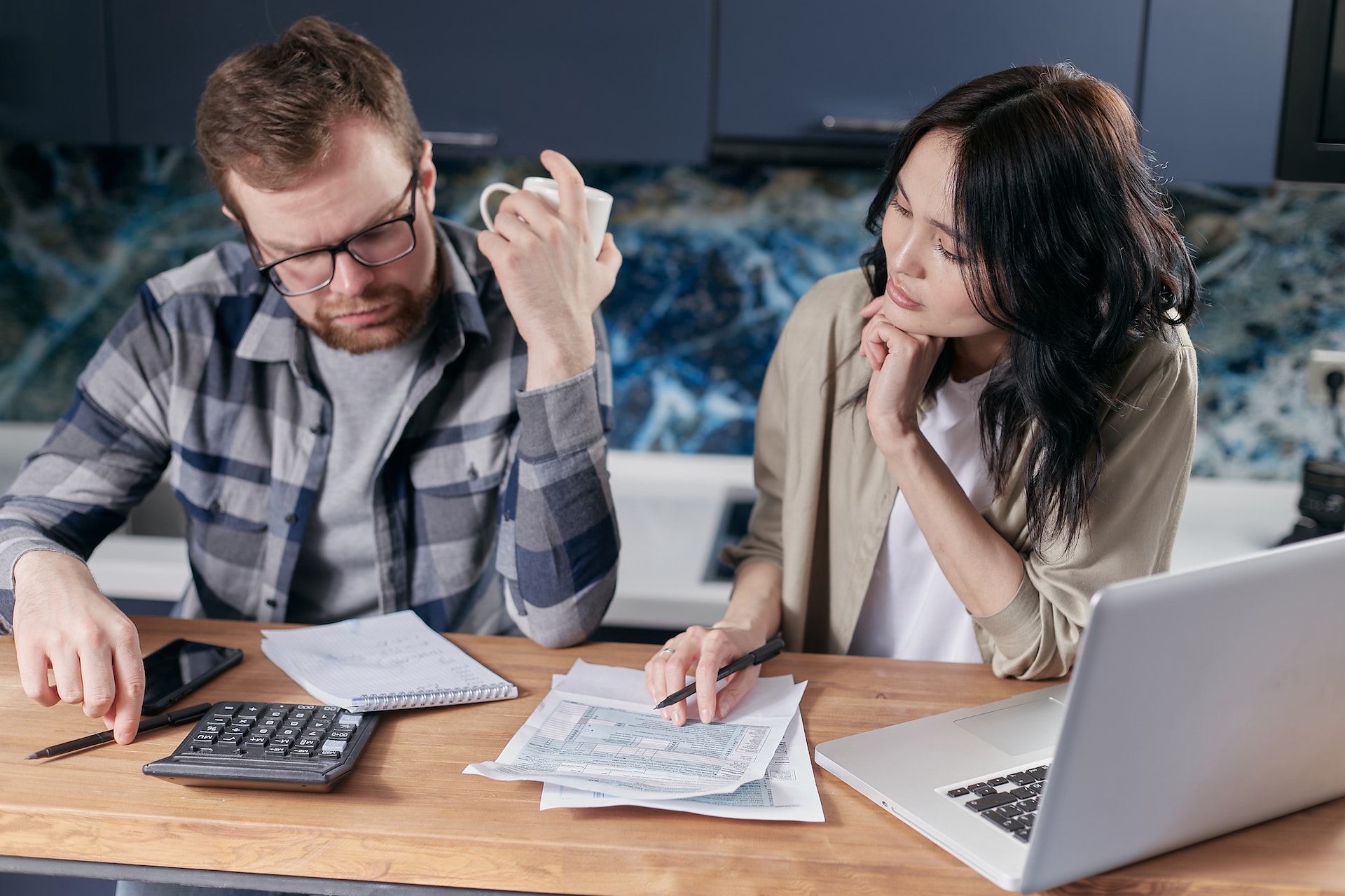 A couple sits at a table in a kitchen, overlooking some documents. There are papers, a calculator, laptop, and notepad on the table. The woman points to a section of a document, while the man holds up a mug and looks down with a pensive expression.