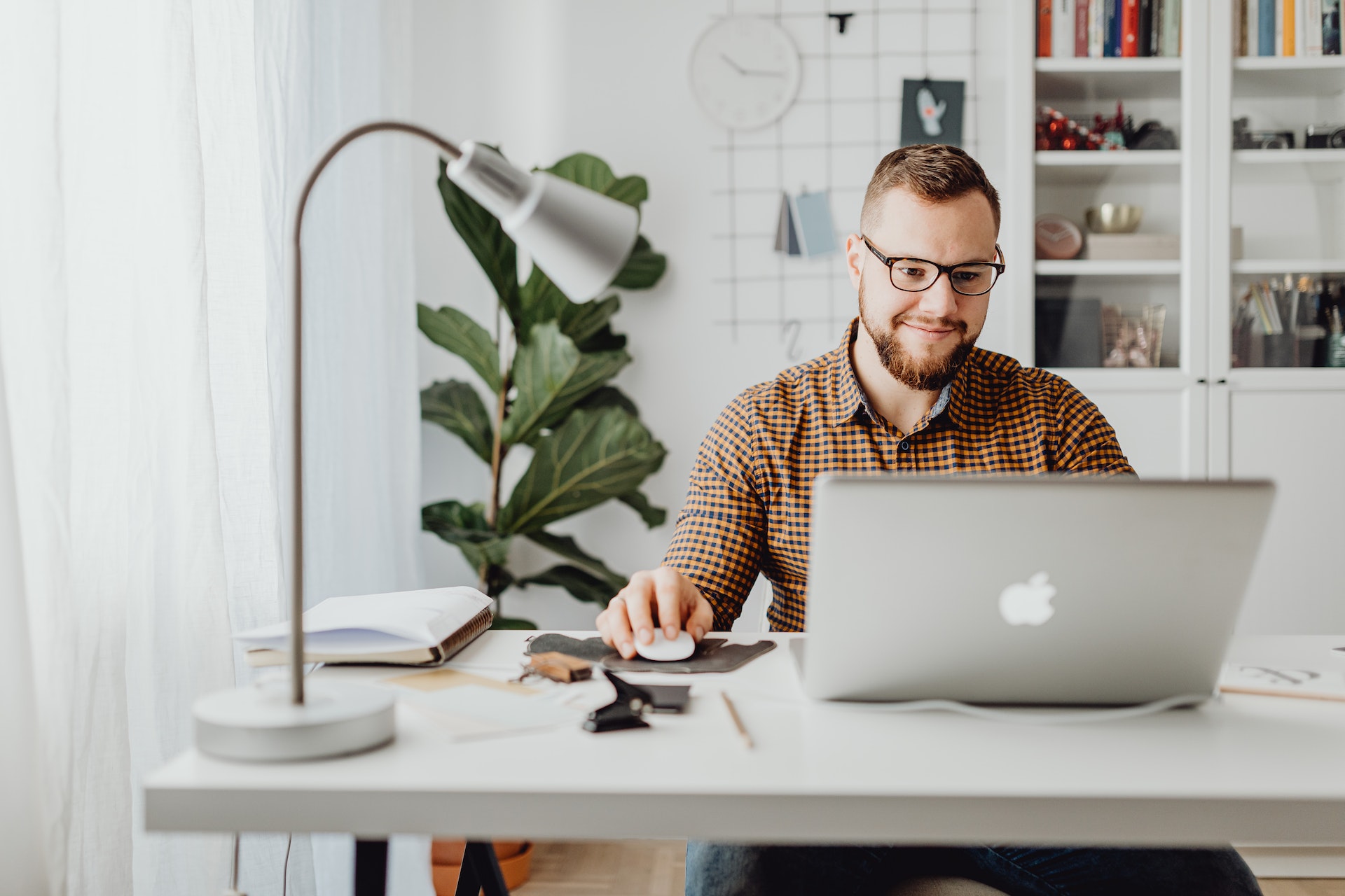 A man with glasses and a high and tight haircut smiles as he browses on his laptop while seated at a desk. There is a small lamp on the desk, as well as notekeeping tools. Behind the man is a bookcase with knickknacks and books. A large potted plant is next to the bookcase.