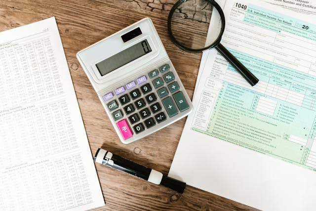 A photo of tax forms, a calculator, dry erase marker and a calculator on a wooden desk.
