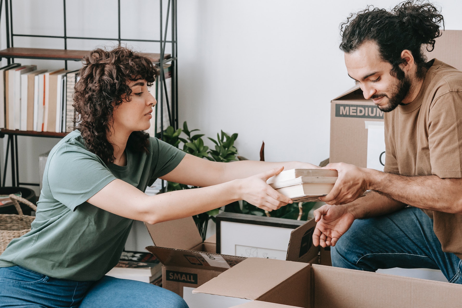 A man and woman sitting on the floor, packing books into open cardboard boxes. A half empty shelf sits against the wall behind them, with a medium sized house plant on the floor beside it. The people are wearing jeans and earth tone tee shirts. Both people have shaggy brown curly hair.