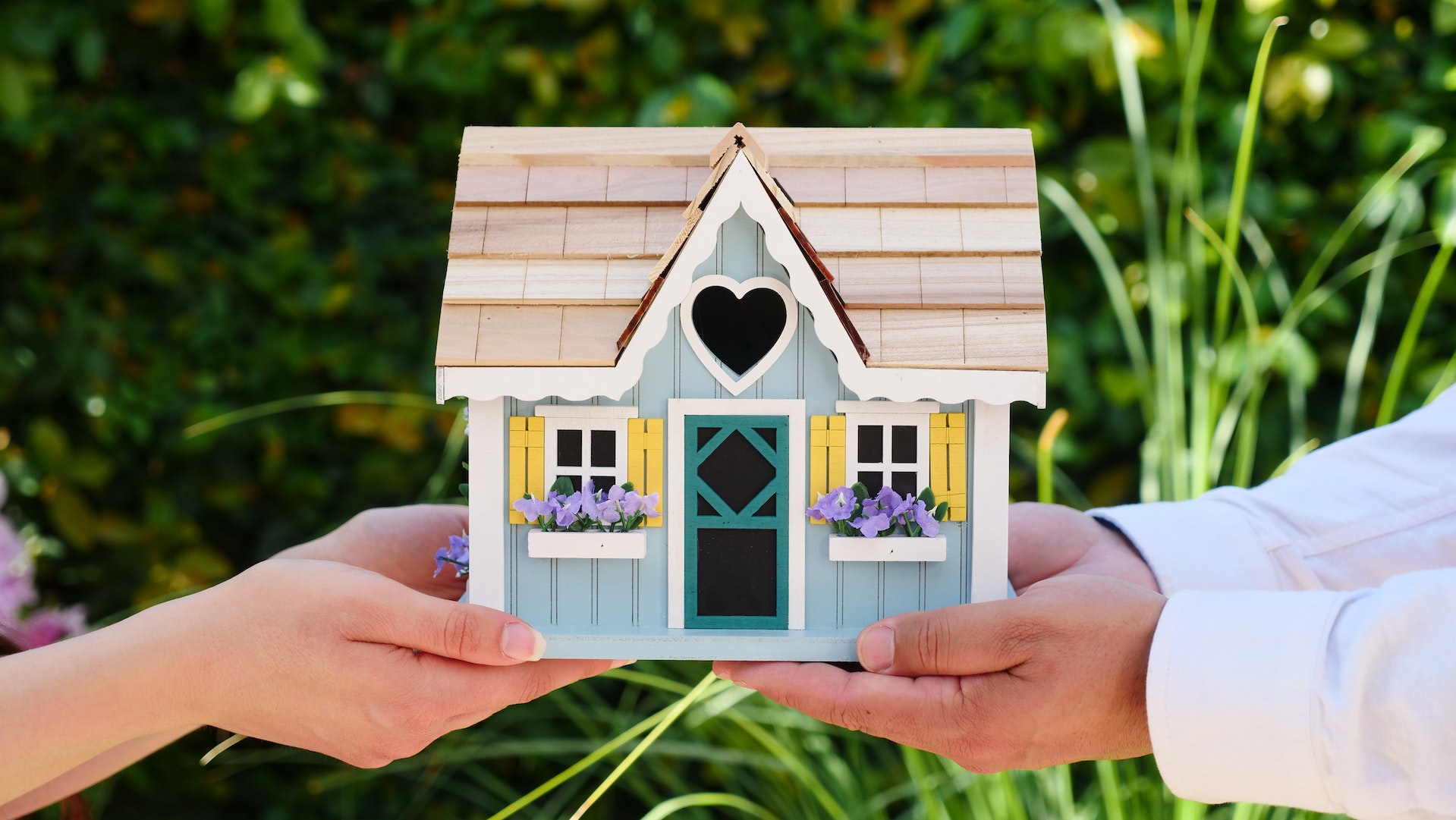 A small-scale model home being supported by four hands, two on each side of the photo. The background is lush green foliage.