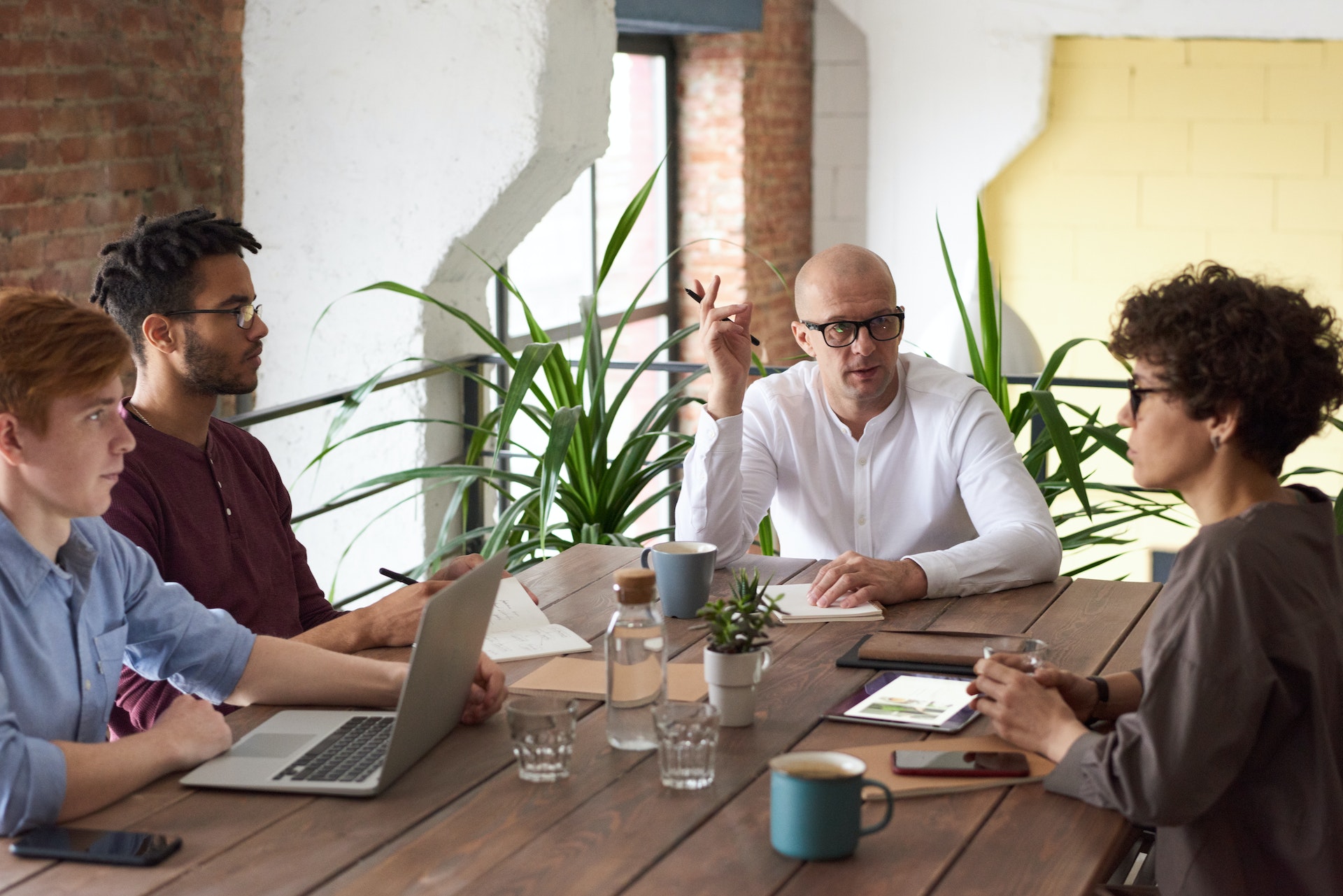 A group of four people sit around a wooden table. A bald man in a white henley shirt wearing glasses sits at the head of the table. There are potted plants behind him. On the right side of the table sits a person with short curly hair, glasses, and a long sleeve brown shirt. Across from them are two young men. One in a blue button down shirt and the other in a maroon long sleeve polo shirt. They appear to be discussing business as there is a laptop, notebooks, cups, mugs, and tablets on the table.