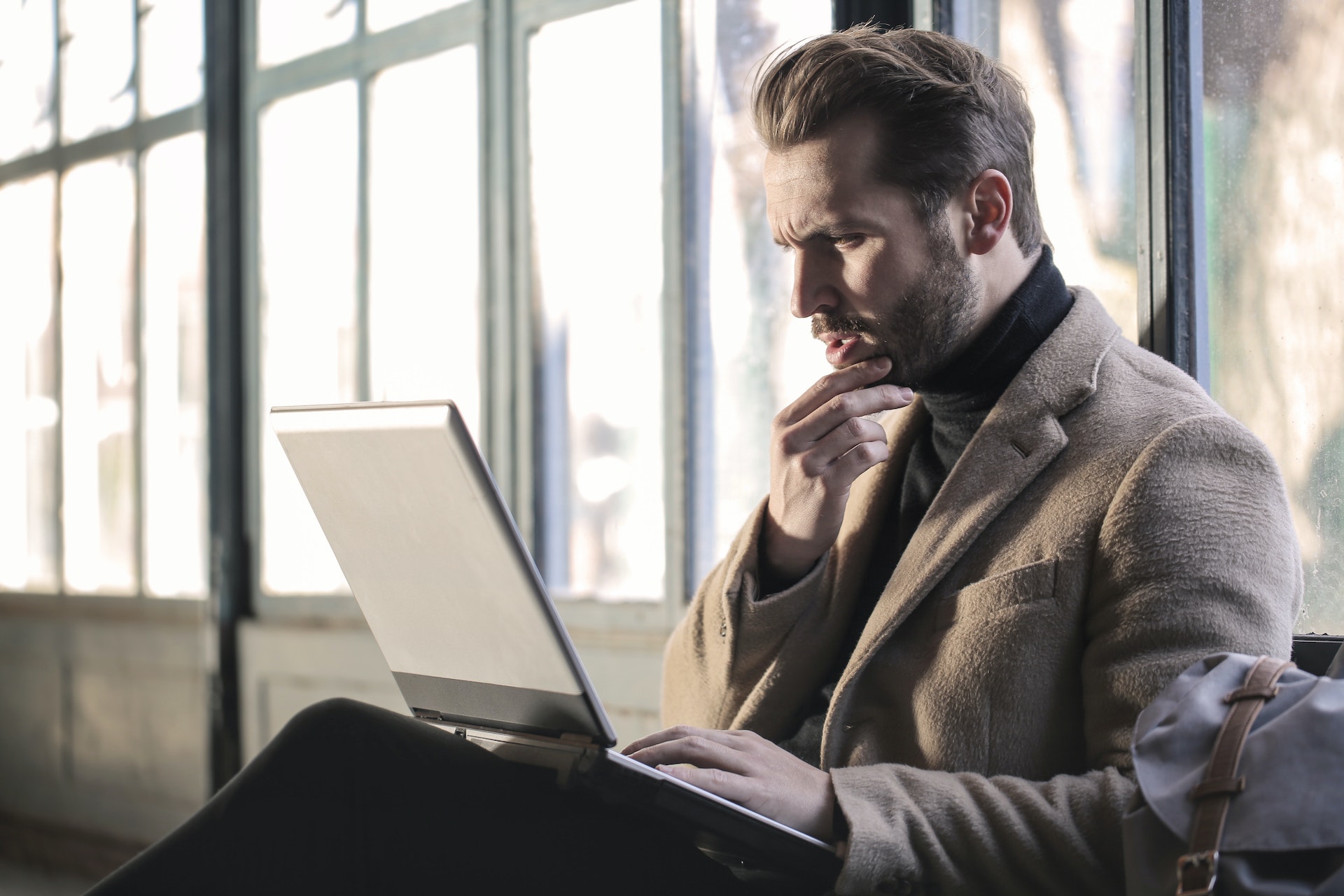 A man in a black turtleneck and earth tone blazer sits against a wall of windows. A laptop is in his lap. He is looking at the laptop with a furrowed brow, one hand on his bearded chin.