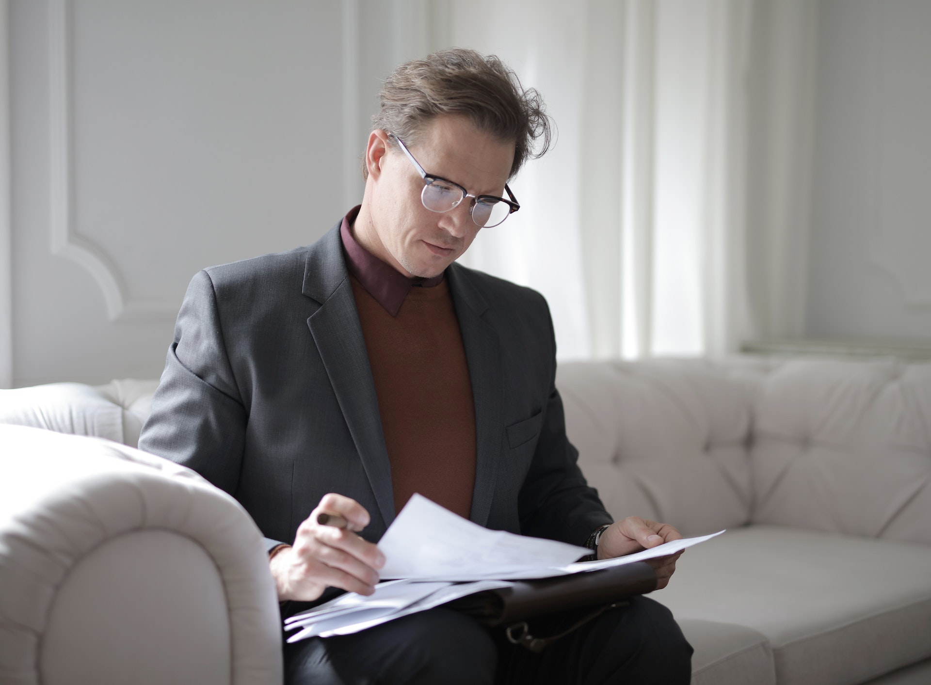 A man in a brown sweater vest, maroon dress shirt, and gray blazer sits on a white couch. He is also wearing glasses, and has short messy brown hair. He looks down at a loose stack of papers in his lap, holding one page in each hand.