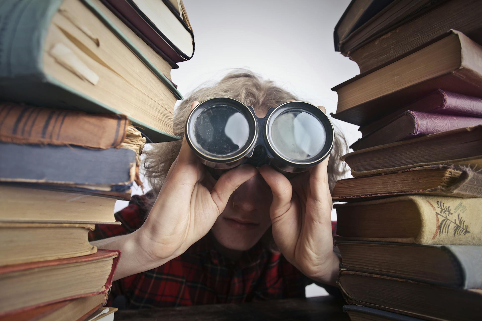 A person peers through binoculars at the camera between stacks of old books.