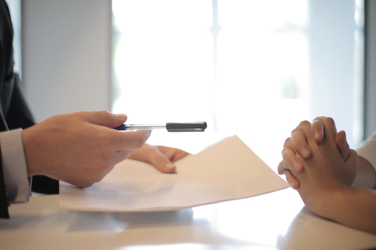 A pair of hands rest on a desk clasped together on the right side of the photo. On the left, another pair of hands offers a pen and paper to their counterpart. A bright window makes up the background of the photo.