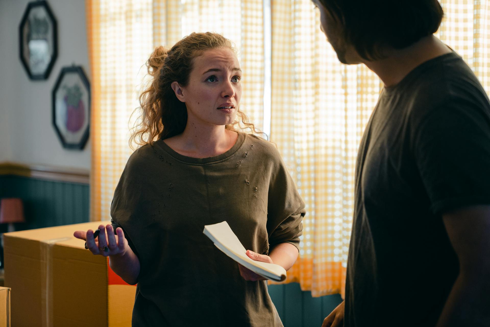 Two people standing in living room with gloomy lighting. There are two octagonal frames on the wall, moving boxes, and yellow checked curtains pulled closed over a bright window. A man is mostly silhouetted and turned to look at a woman in focus. The woman wears a muddy green sweatshirt and has wavy light brown hair that is loosely tied back. She holds a stack of papers in one hand and the other hand is raised in a questioning manner. She has an expression of bewilderment and upset.