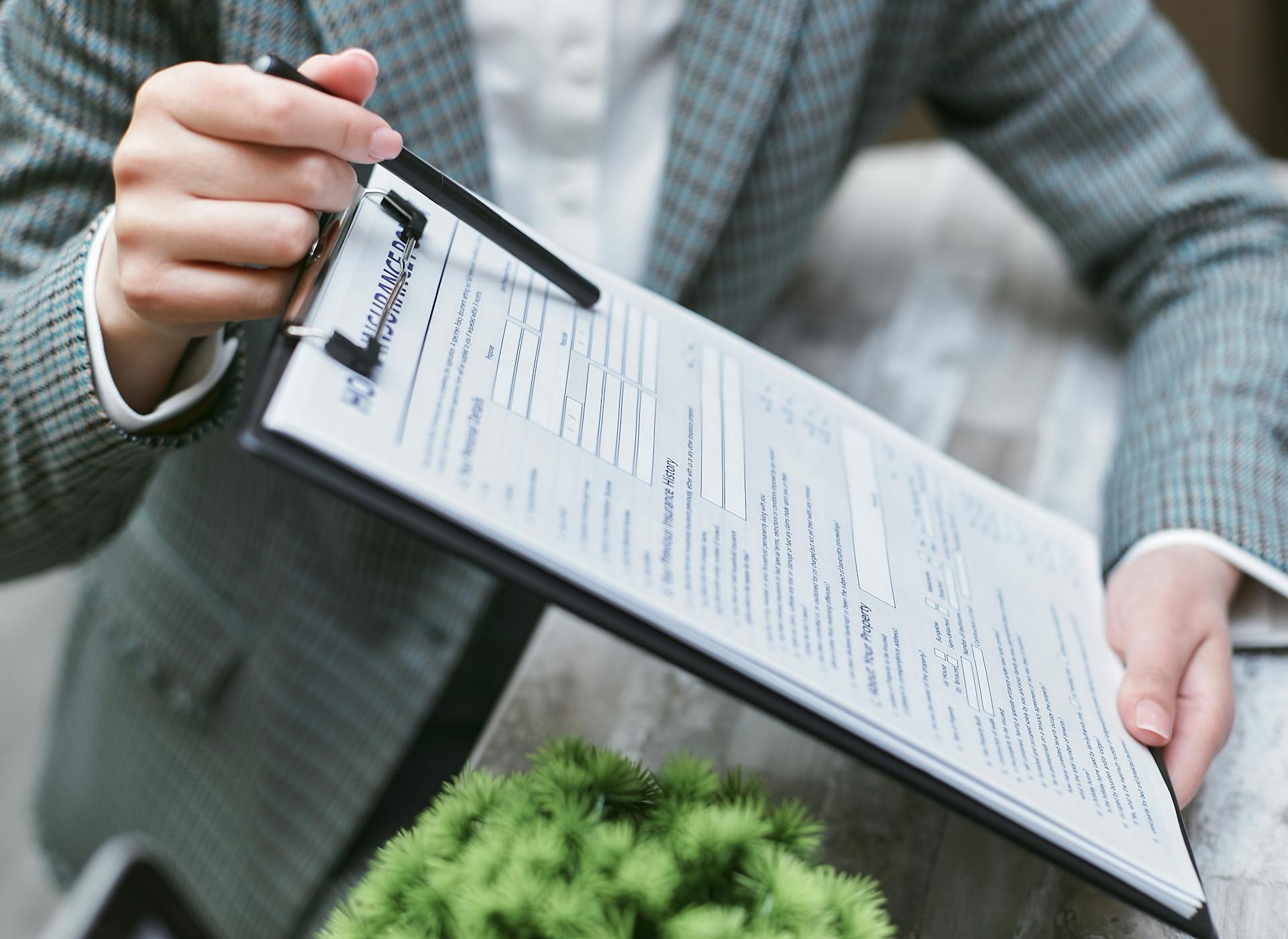 A person in a pistachio and brown plaid blazer holds a clipboard with an insurance form on it. In one hand, they hold a pen and point to a blank section of the form.